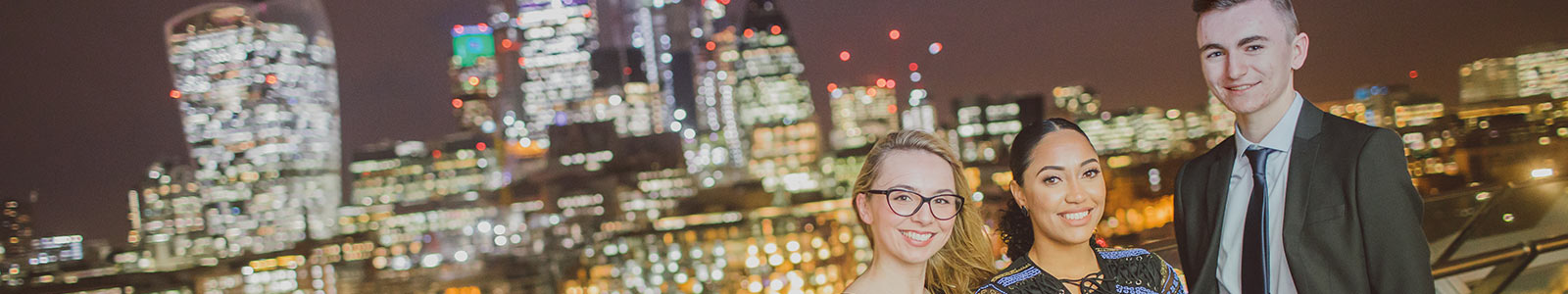 Three students smiling in front of a city in lights background