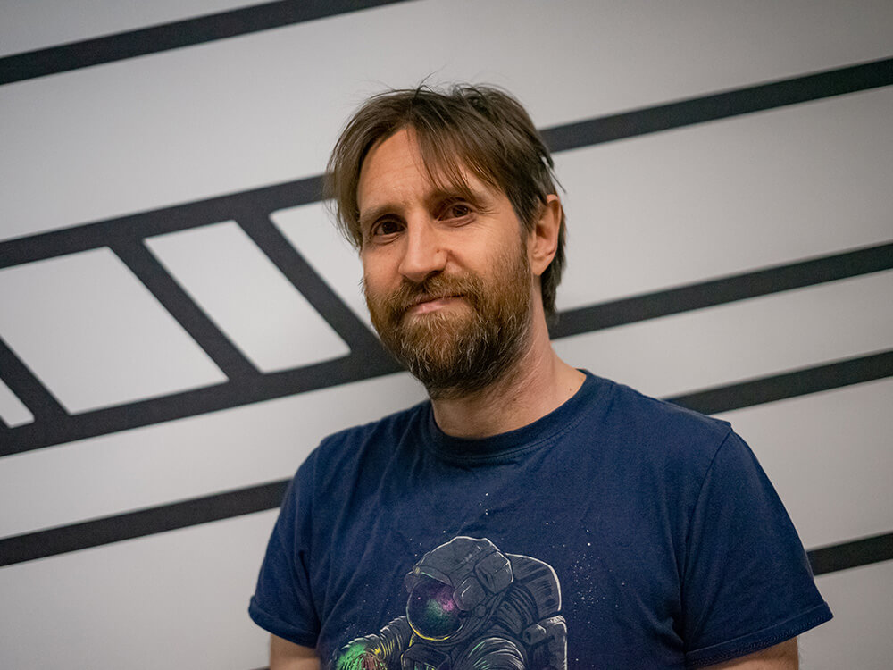 Headshot of a white man with beard and navy t-shirt and facing camera with white and black striped walls in background
