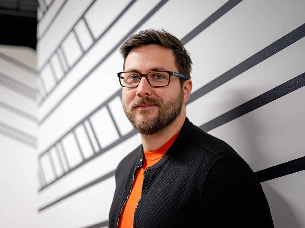 A white man with a beard and glasses, orange t-shirt and black shirt standing against a white wall with black stripes