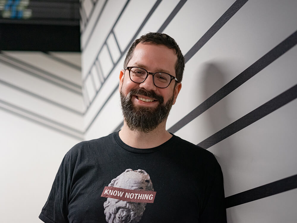 Headshot of a white man wearing glasses, a t-shirt and a beard and facing camera with white and black striped walls in background